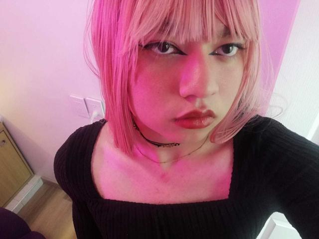 OliverPelman with pink hair, black ribbed shirt, choker, and pink lighting, has full lips and a serious expression, showing a cute face with medium tits.