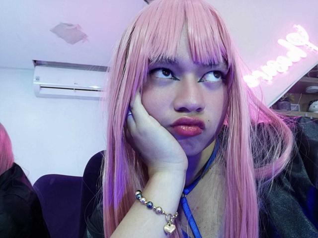 OliverPelman with pink hair, pouty lips, and blue eyes, hand on cheek, wearing a blue necklace and bead bracelet, looking sideways. Purple and blue lighting.