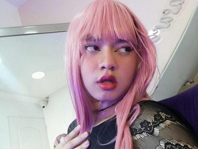 OliverPelman with pink hair, wearing black lace top, has shiny red lips, looking to the side, hand near neck, in a brightly lit room.