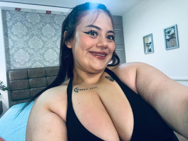 EllaRider with large tits in a black halter top, smiling, has tattoos on her chest, long brown hair, and black eyeliner, standing in a bright room.