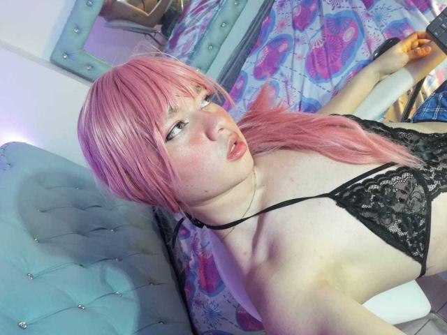 OliverPelman with pink hair, wearing black lace bra, lying on blue tufted bed, holding black high heel, pale skin, pink lips, looking up with slightly parted mouth.
