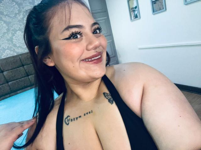EllaRider with dark brown hair, black eyeliner, and smile, wearing a black halter top, showing large tits and a tattoo above her cleavage. Her hand touches her hair.