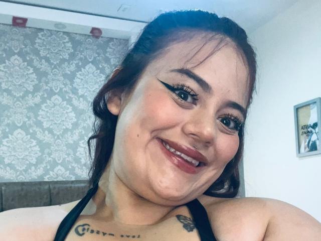 EllaRider with dark brown hair, bold black eyeliner, smiling, showing her nose ring, black halter top, tattoos on her neck and chest, and cute freckles.