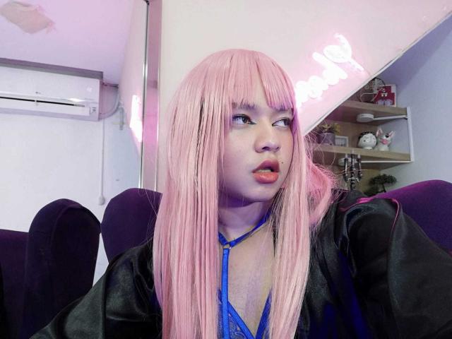 OliverPelman with pink wig, blue lace-up top, black jacket, purple chair, and air-conditioner in background, looking to the side with pouty lips and slightly parted lips.
