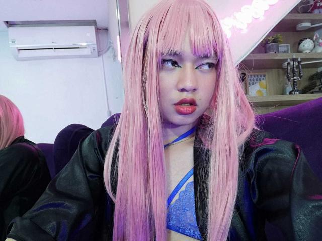 OliverPelman with pink hair, blue lace bra, and black jacket, has red lips and a slightly open mouth, sitting on a purple couch, looking to the side.