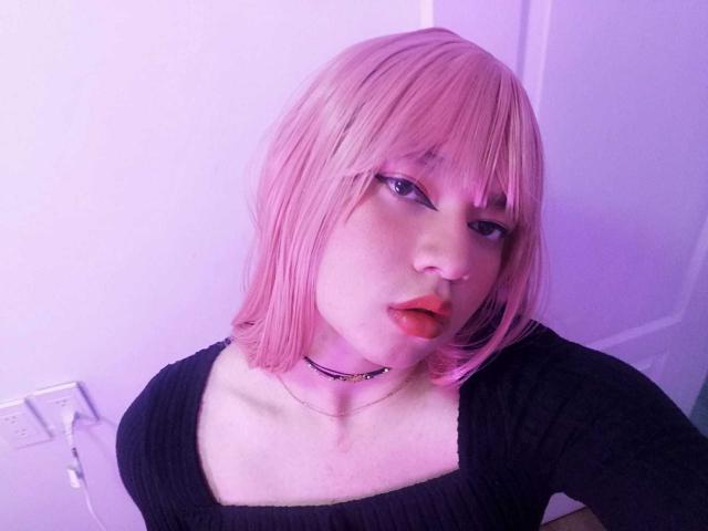 OliverPelman has pink bob hair, black top, red lips, and a black choker, with a serious expression, in purple-lit room. Boobs visible under top.