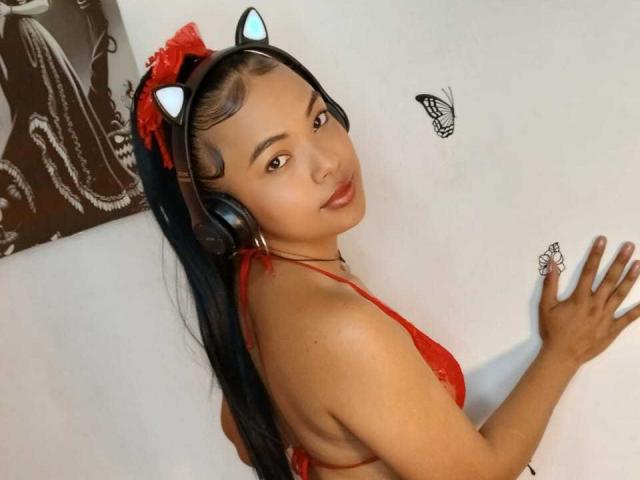 SoleneFontaine with black hair in a ponytail, wearing a red lace bra, cat ears, headphones, and a red bow, looking back with a butterfly on the white wall.