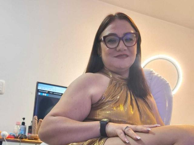 LadyNylon, avec des lunettes, une robe dorée métallique et un bracelet noir, assise avec les fesses légèrement relevées, souriante. Elle a une poitrine de taille moyenne, une peau claire et des cheveux bruns..