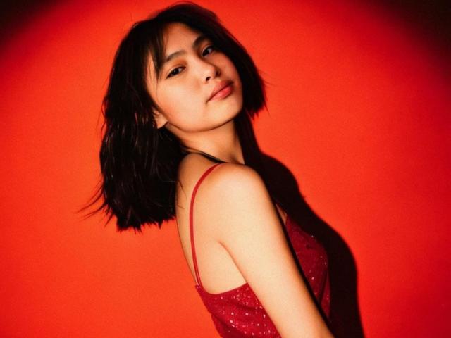 Jennelyn, avec ses cheveux noirs courts, portant une robe rouge scintillante, montrant sa poitrine et ses fesses, regardant l'appareil photo avec un léger sourire, sur un fond rouge vif..