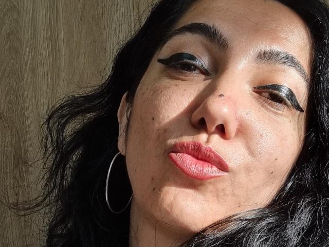 Le visage de SexyMirra, avec ses cheveux noirs ondulés, son trait d'eye-liner noir épais, son rouge à lèvres rouge, son petit nez, ses taches de rousseur, ses boucles d'oreilles argentées, sa peau légèrement brune, ses lèvres légèrement charnues, éclairé par le soleil, avec un fond en bois..