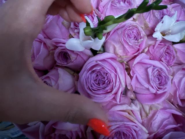 Didiene's hands with red and orange nails are touching a bunch of pink roses and white flowers, removing a green stem. Her fingers are slightly curled.