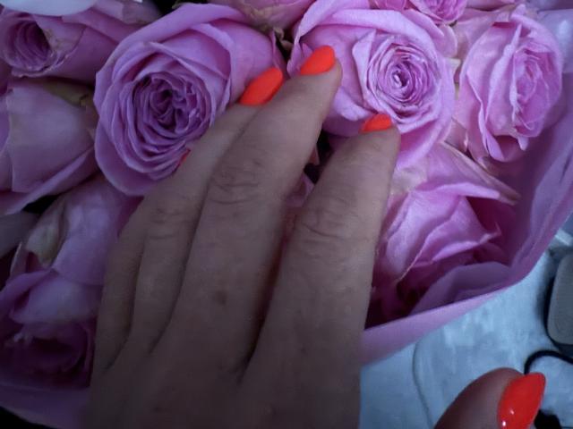 Didiene's hand with bright red nails touches a bunch of pink roses. Her fingers are slightly curled, and the roses are fully bloomed.