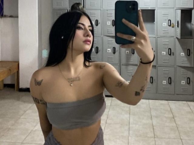 NatallieBlue with black hair, tattoos, and gray tube top, holding phone, showing tits, in locker room, taking selfie, looking at mirror.