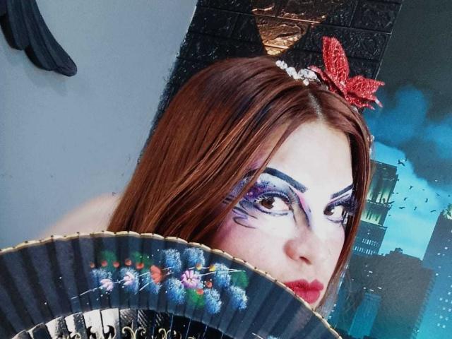 SkylarDesire with red hair, wearing a red butterfly headpiece, bold purple eye makeup, and holding a black fan with colorful patterns, has pale skin and red lipstick.