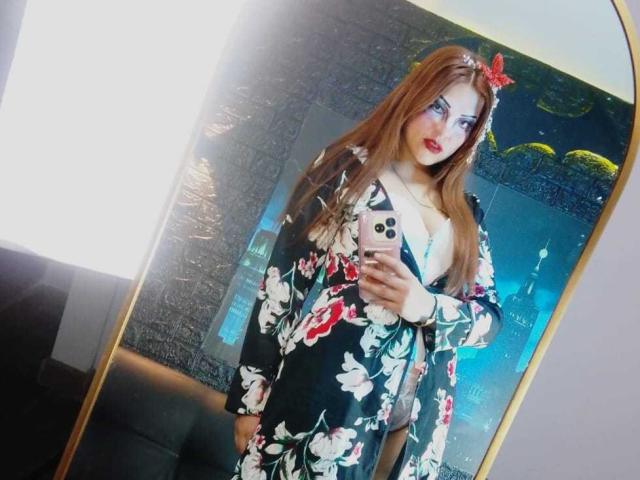 SkylarDesire with long brown hair, floral black dress, red lipstick, blue eyeshadow, and red butterfly hairclip, holding phone, reflecting in mirror, showing medium tits and slim build.