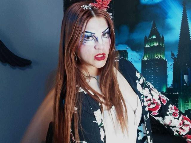 SkylarDesire has long brown hair, white makeup with blue eyeshadow, red lips, floral jacket, and black floral pattern, looking at camera with serious expression, wearing red headpiece.
