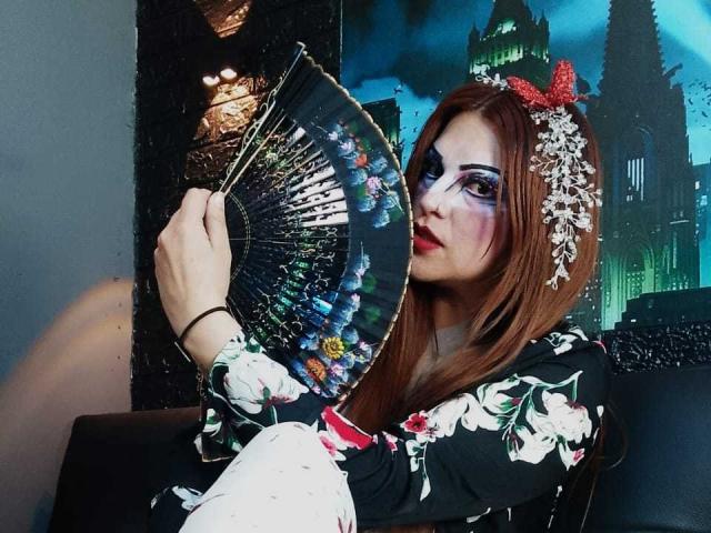 SkylarDesire with long brown hair, floral kimono, and white gloves, holding a colorful fan, wearing purple eye makeup, red bow, and floral headpiece, with a serious expression.