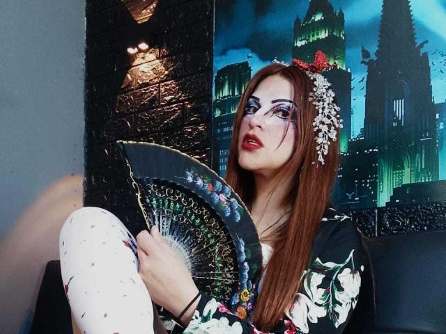 SkylarDesire with long brown hair, floral headpiece, and black floral dress, holding a decorative fan, wearing white patterned tights, sitting, looking up, red lipstick.