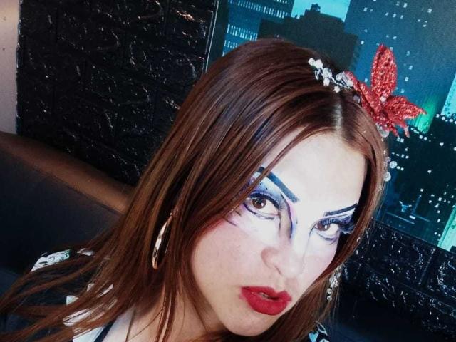 SkylarDesire has red lipstick, blue and white dramatic eye makeup, brown hair with a red glitter butterfly headband, and hoop earrings. Her tits are partially visible.
