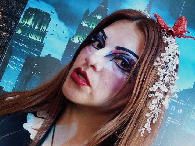 SkylarDesire with dark, dramatic eye makeup, red lipstick, white floral headband with red bow, brown hair, and blue-eyed look, wearing black and white outfit.