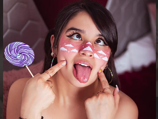 MiaNickole with face painted white and pink clouds, holding a purple swirl lollipop, tongue out, and pointing with fingers at her cheeks, showing off her colorful nail polish.