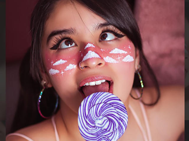 MiaNickole has white cloud makeup on her cheeks, a nose ring, and colorful earrings. She's licking a purple and white swirl lollipop, eyes looking up.
