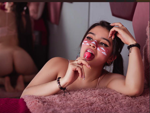 MiaNickole with white face paint, tongue out, hand on head, lying on pink fur, bare breasts, butt in mirror reflection, black bracelets, dark hair, pink room.