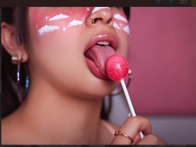 MiaNickole's face with pink face paint, nose ring, and hoop earrings, licking a red, grid-patterned lollipop, her tongue out, wearing a pink hoop earring on her right finger.