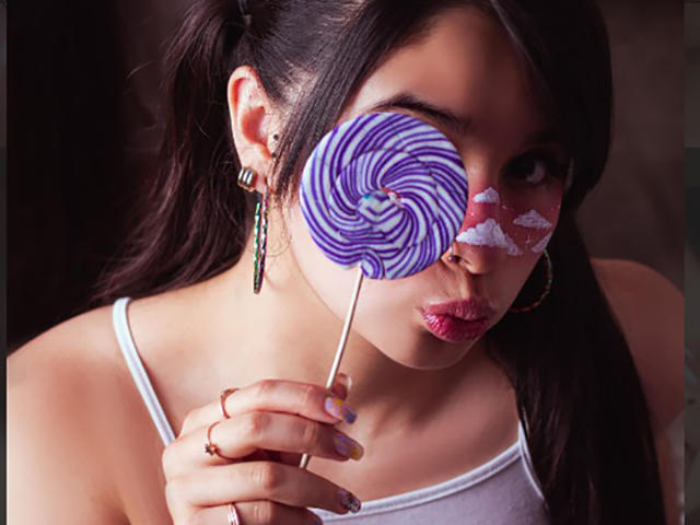 MiaNickole with purple swirled lollipop in face, white candy patterns on cheek, pouty lips, dark ponytail, white tank top, hoop earrings, and a ring on her finger.