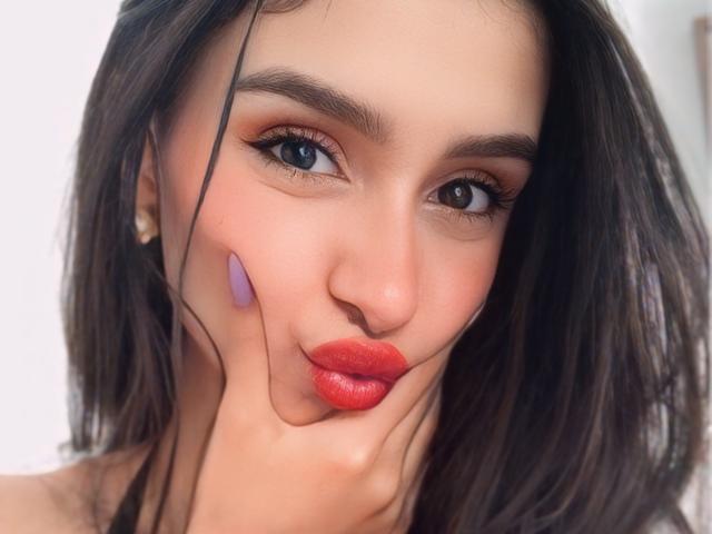 CamilaJolie has red lipstick, purple makeup on her cheek, dark eyes, and a slight pout, with dark hair and pearl earrings, looking sexy and cute.