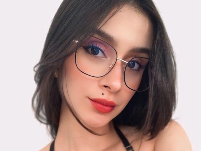 CamilaJolie with red lips, dark straight hair, purple eyeshadow, black-rimmed glasses, and a nose piercing, looking at the camera with a slight smile. Boobs not visible.