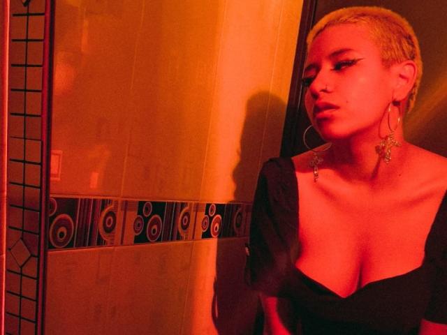 DulceSweett with short blonde hair, wearing a black dress with puffed sleeves, large earrings, and red lighting, showing off her tits and a confident, sexy expression.