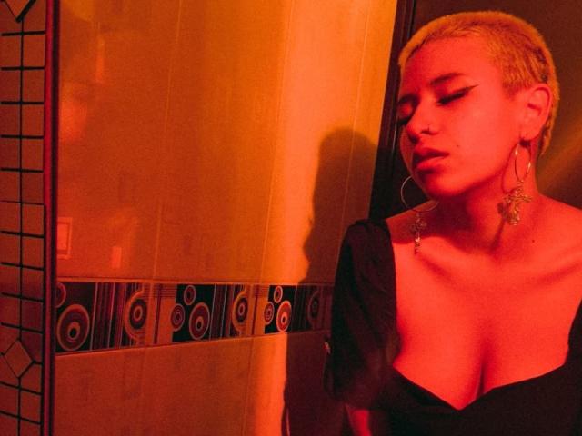 DulceSweett with short blonde hair, red lighting, closed eyes, black dress, cleavage, large hoop earrings, and nose ring, standing against a wall with a shadow.