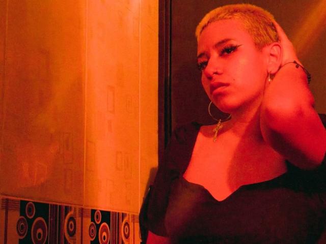 DulceSweett, com cabelo loiro curto, brincos grandes de argola, blusa preta e piercing no nariz, em pé sob uma luz laranja quente, com a mão no cabelo, com uma expressão ligeiramente irritada, com seios visíveis..