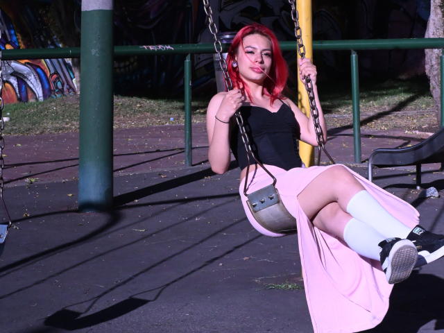 RouseParker with red hair, black top, pink skirt, white socks, black shoes, sitting on a swing, holding chains, looking at camera, sunlight, playground.