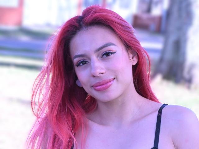 RouseParker with bright red hair, smiling with pink lipstick, black strap dress, and subtle eyeliner, has a cute face and small tits, standing in a sunny outdoor setting.