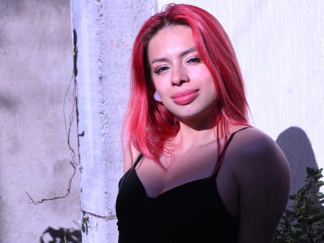 RouseParker with red hair, wearing a black spaghetti-strap top, pouty pink lips, and white earbuds, standing against a sunlit, textured wall, showing her small tits.