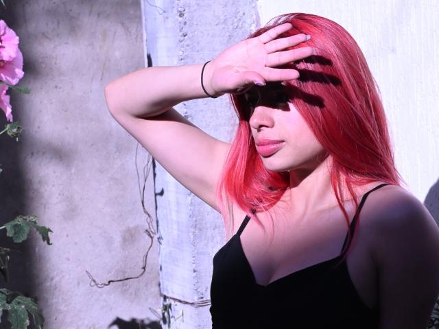 RouseParker with red hair, black spaghetti top, and pink lips, shielding eyes with right hand, wearing a black bracelet, in bright sunlight.