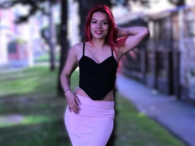 RouseParker with red hair, black strapless top, and pink skirt, standing with one hand on her head, smiling. She has medium tits and a cute, sexy look.