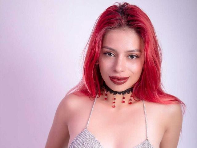 RouseParker with red hair, wearing a silver halter top, black choker with red beads, and dark lipstick, smiling at the camera. Boobs are small and her ass is not visible.