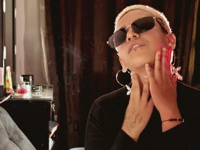 DulceSweett with short blonde hair, wearing dark sunglasses, large hoop earrings, black top, and tattoo on hand, resting cheek on hand, looking up with a slight smile.
