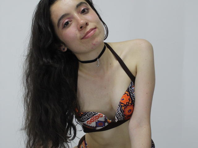 ElodiePerrine with long, wavy black hair, wearing a colorful bra with orange, black, and white patterns, black choker, and smiling slightly, showing small, perky tits.