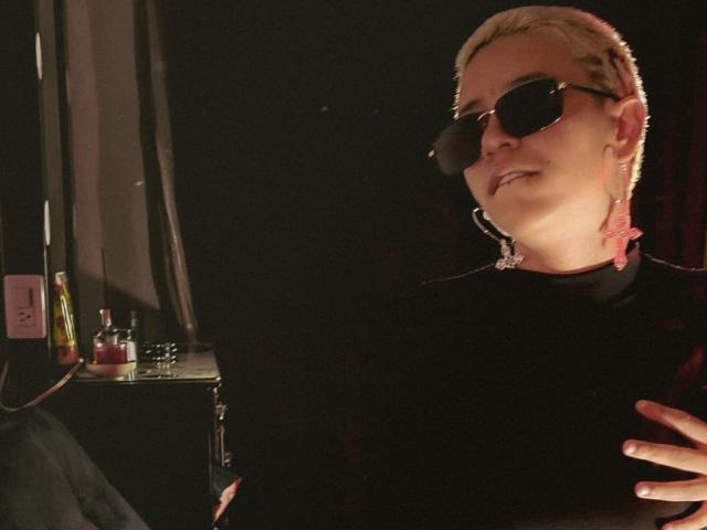 DulceSweett, avec ses courts cheveux blonds, ses lunettes de soleil et ses piercings roses aux oreilles, portant un haut noir, debout dans une lumière tamisée, une main sur la poitrine, un léger sourire..