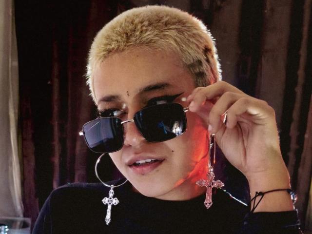 DulceSweett with short blonde hair, wearing black sunglasses, large hoop earrings, and pink cross earrings, holding a pink cross earring near her face. Her lips are slightly parted.