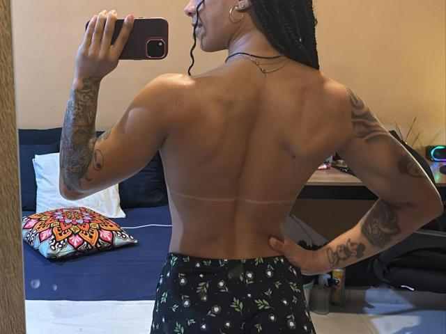 ManuHot69 with tattoos, muscular back, braided hair, tan lines, and black patterned shorts, taking a mirror selfie, holding a red phone.