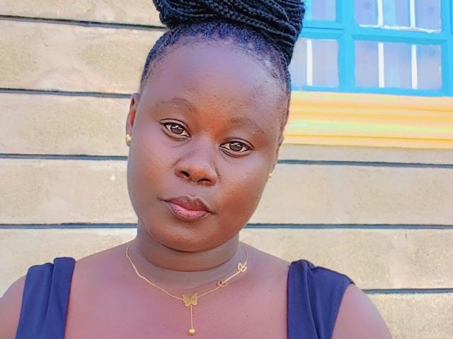 StilletoBabe with dark brown skin, short braided hair in a bun, gold necklace, small gold earrings, pouty lips, wearing a black sleeveless top, standing against a wall.