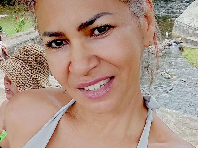 BrigiteWalsh with a smile, showing her pink lipstick and brown eyes, wearing a gray halter top, and her blonde hair in a ponytail, with a man in a hat behind.
