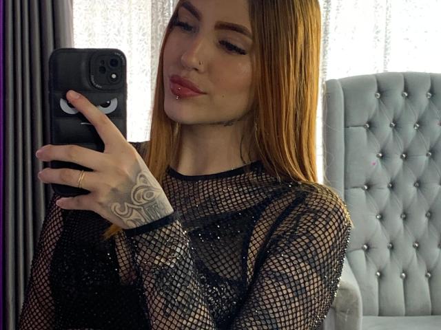 Antonella Brith, avec ses longs cheveux roux, portant un haut en filet noir, prend un selfie devant un miroir avec son téléphone, montrant un tatouage sur sa main droite et faisant la moue avec ses lèvres roses..