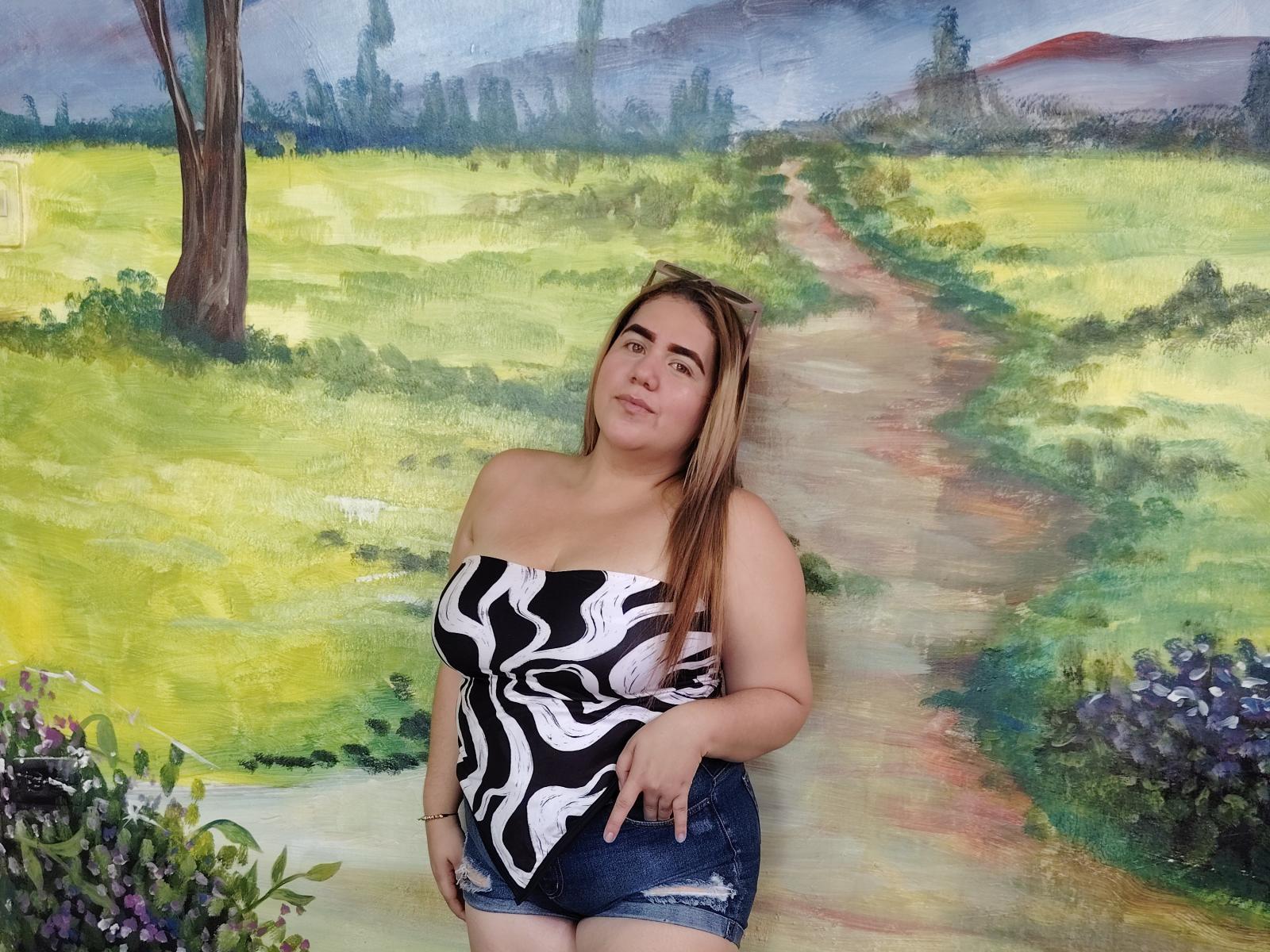 CloeGreco, with medium tits, wears a black-and-white wavy top and denim shorts, standing against a painted countryside, looking slightly up with a relaxed expression.
