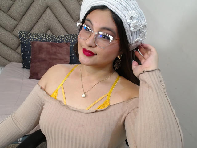 HelueJamil with glasses, red lipstick, beige off-shoulder top, yellow bra straps, headscarf, hand in hair, gold necklace, and earrings, smiling. Her tits are visible through the top.